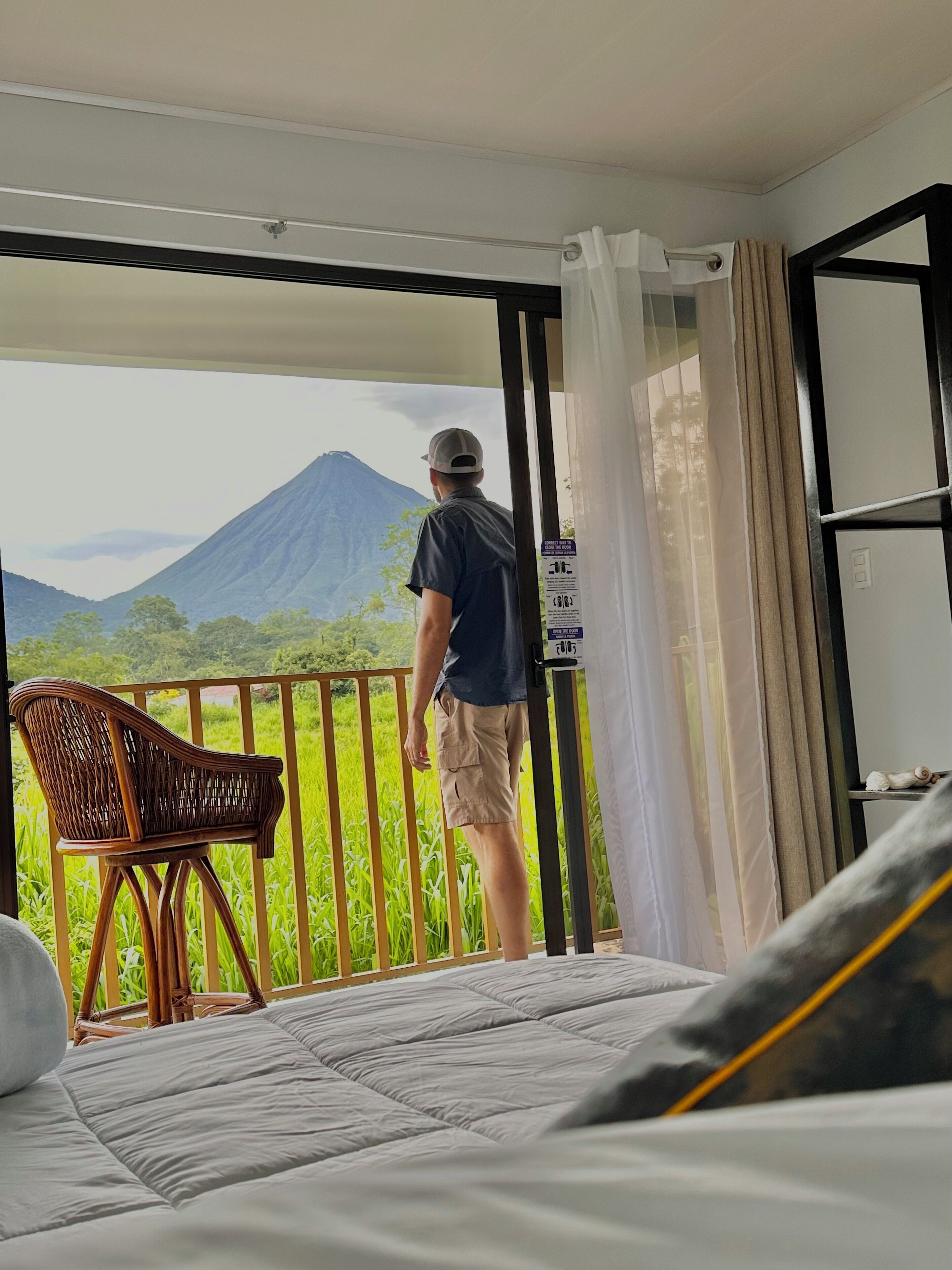 A room with a bed, a wicker chair, and a view of a mountain.