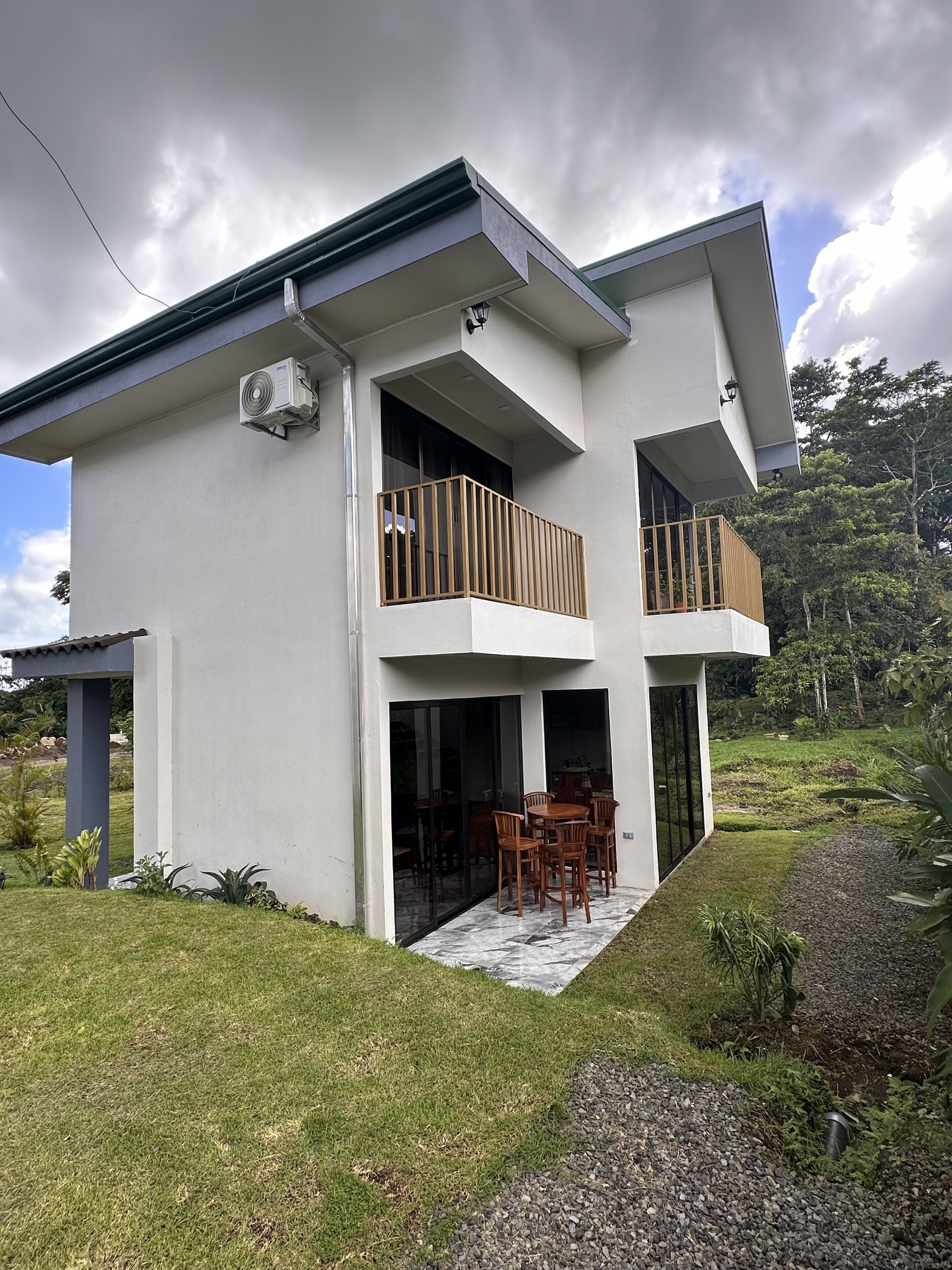 Una casa moderna de dos pisos con balcón, un patio con mobiliario exterior y un jardín.