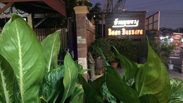 Une enseigne pour un restaurant appelé « Baan Bussa », avec un néon, entourée de grandes plantes vertes.