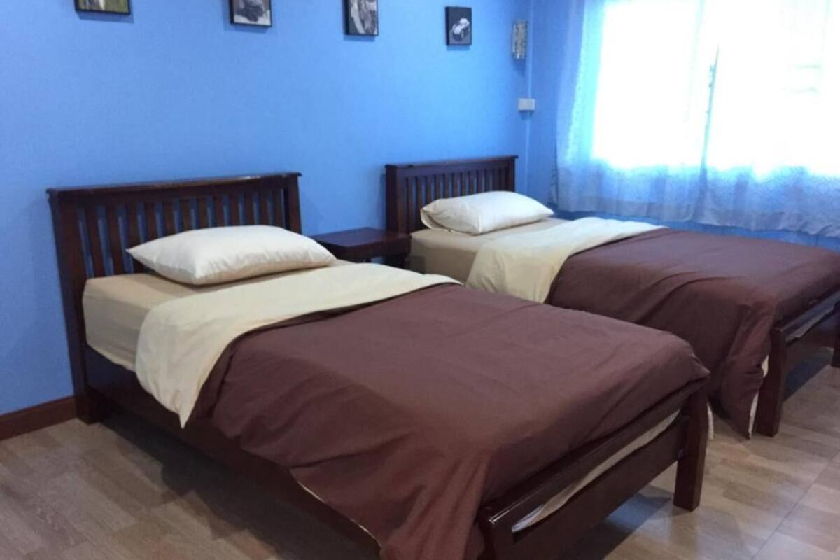 Una habitación con dos camas de madera, cada una con una colcha marrón y un almohadón blanco. Las paredes están pintadas de azul, y hay una ventana con una cortina translúcida.