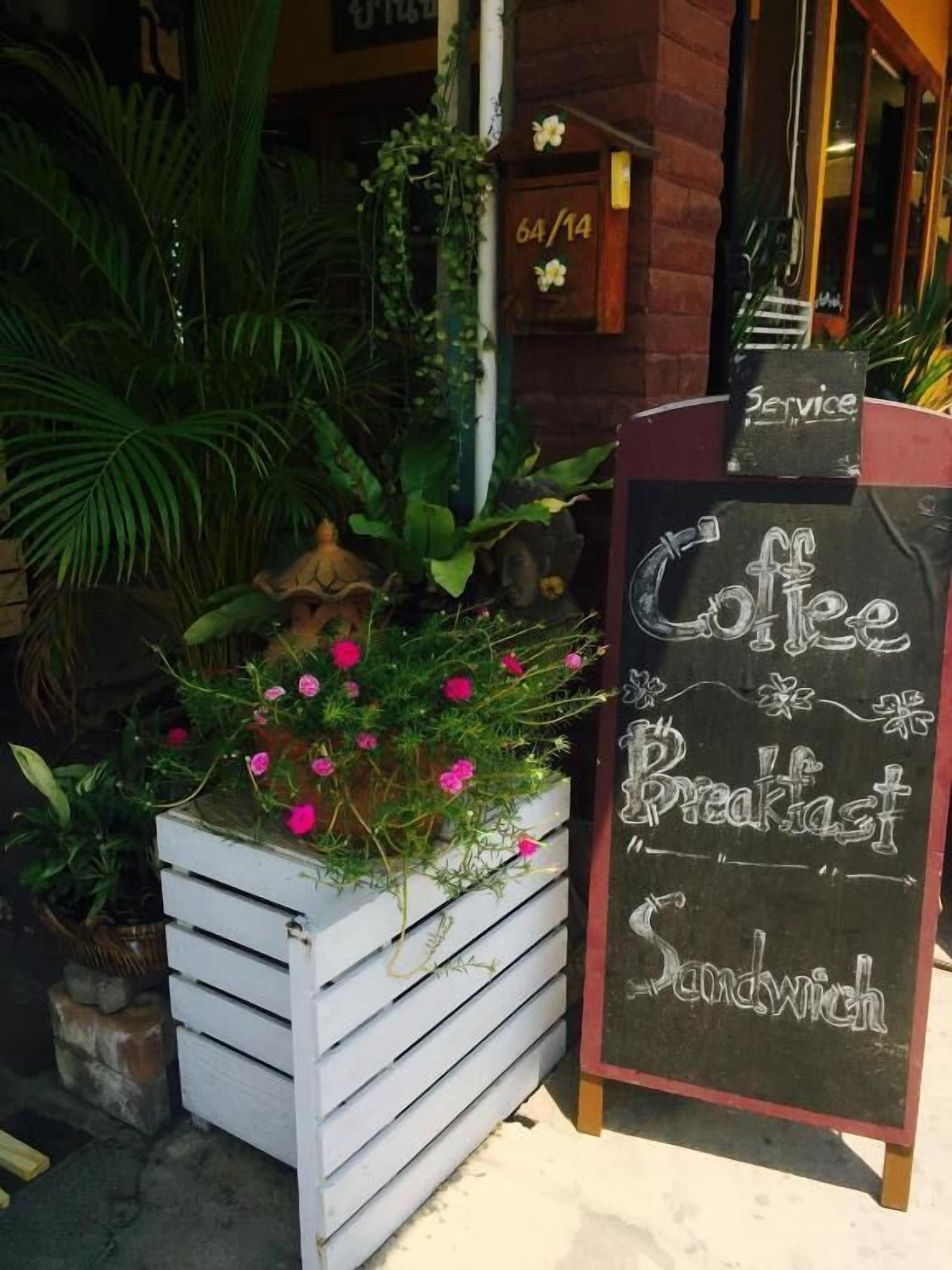 A chalkboard sign advertising coffee, breakfast, and sandwich services outside a building with a brick facade and a number '64/14' above the door.