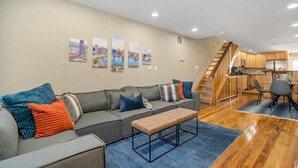 A modern living room with a grey sofa, a wooden staircase, and a kitchen area. - Charming Baltimore Townhome w/ Scenic Views (Baltimore)