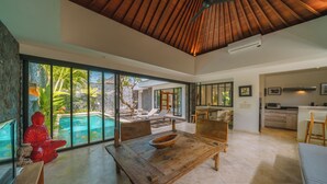 A spacious living area with a wooden dining table, a red Buddha statue, and a large glass window overlooking a pool and tropical garden. - Villa Anahata (Seminyak)
