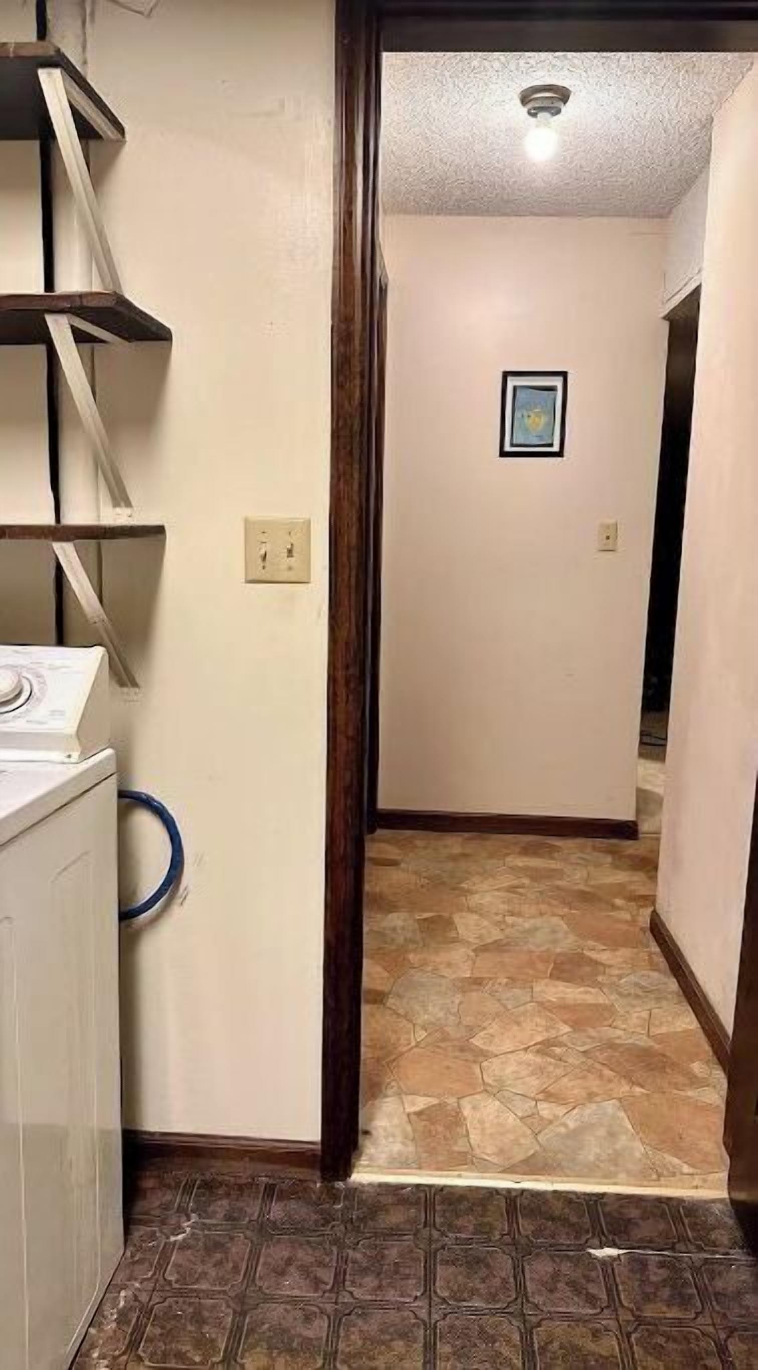 A laundry room with a washing machine, a dryer, and a blue hose. There is a hallway with a tiled floor leading to another room.