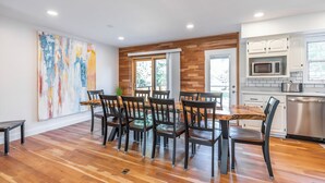 Zona pranzo con tavolo e sedie in legno, un'ampia pittura astratta appesa al muro e una cucina con mobili bianchi ed elettrodomestici in acciaio inossidabile.
