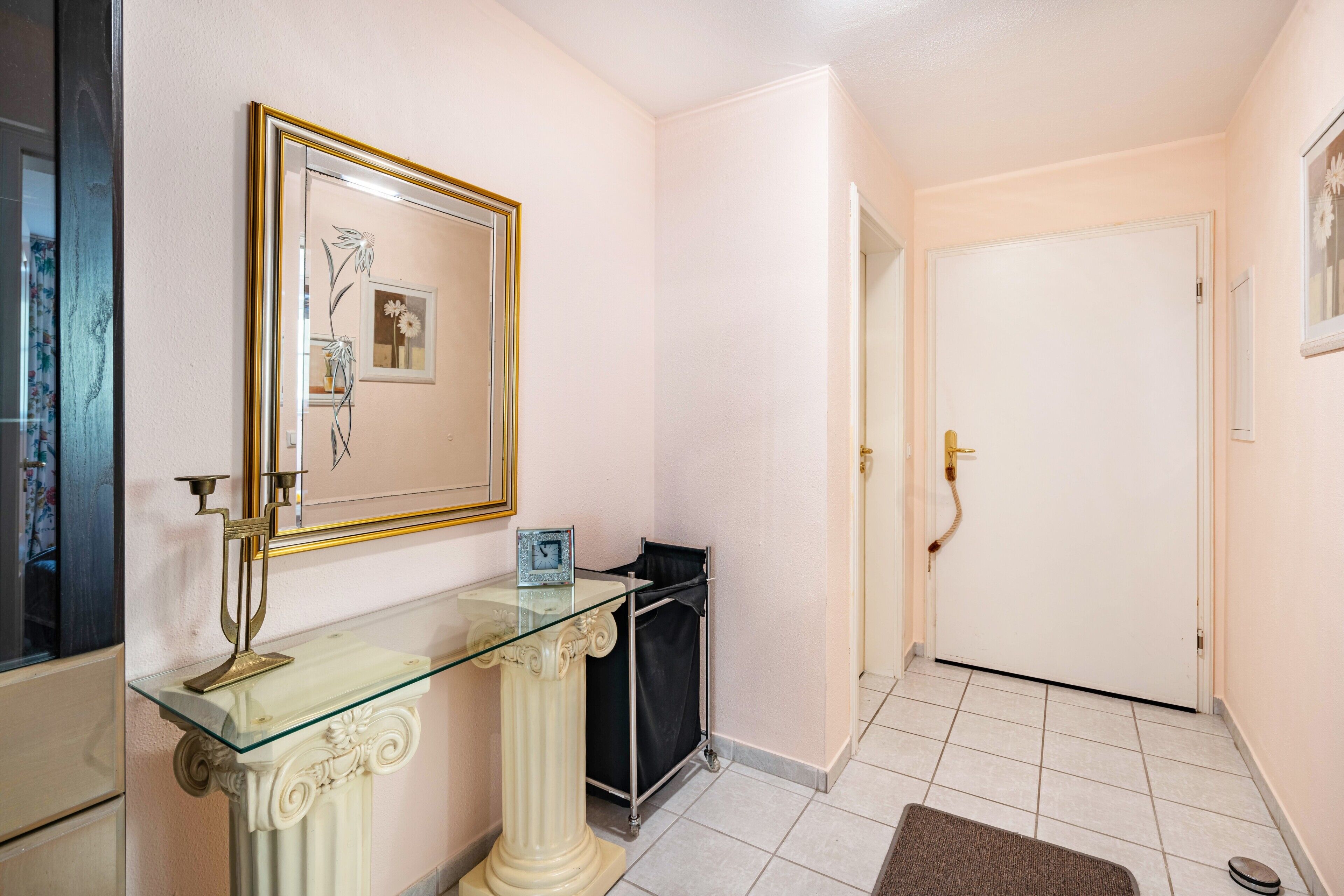 Um corredor com uma mesa consola de vidro, um quadro emoldurado, uma porta e chão revestido de azulejos.