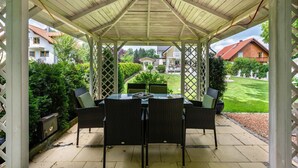 Eine überdachte Terrasse mit Tisch und Stühlen, umgeben von einem Gitterzaun, mit Blick auf einen gepflegten Garten und Wohnhäuser.