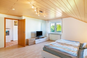Ein Schlafzimmer mit einem Bett, einem Fernseher auf einem Schrank, einem Fenster mit Blick auf Grünflächen und einer Holzbalkendecke.