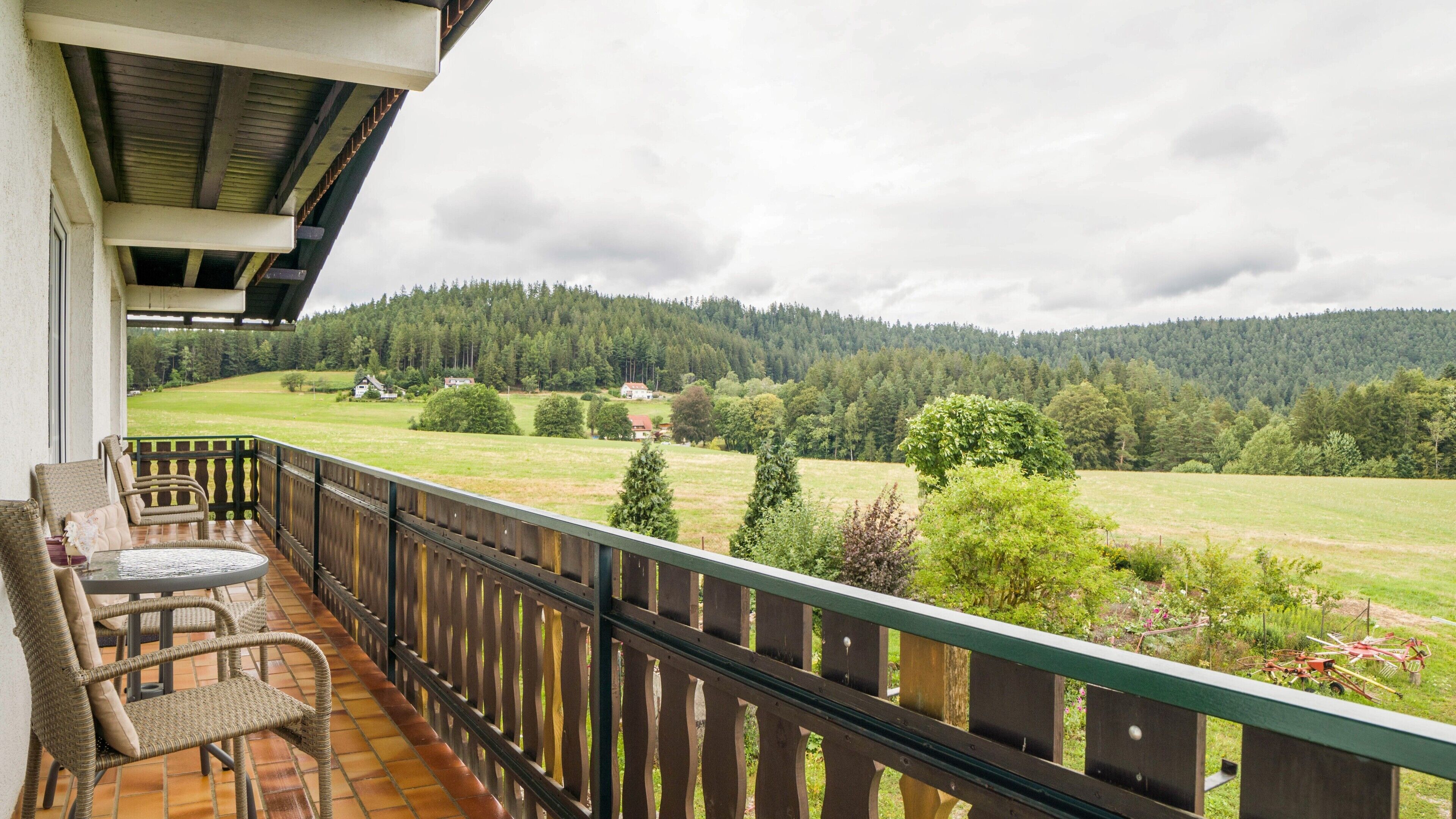Ein Balkon mit Korbmöbeln und Blick auf eine grüne Landschaft mit Bäumen und Häusern.