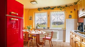 A kitchen with a red refrigerator, wooden chairs, a dining table, and a view of the countryside through two windows.
