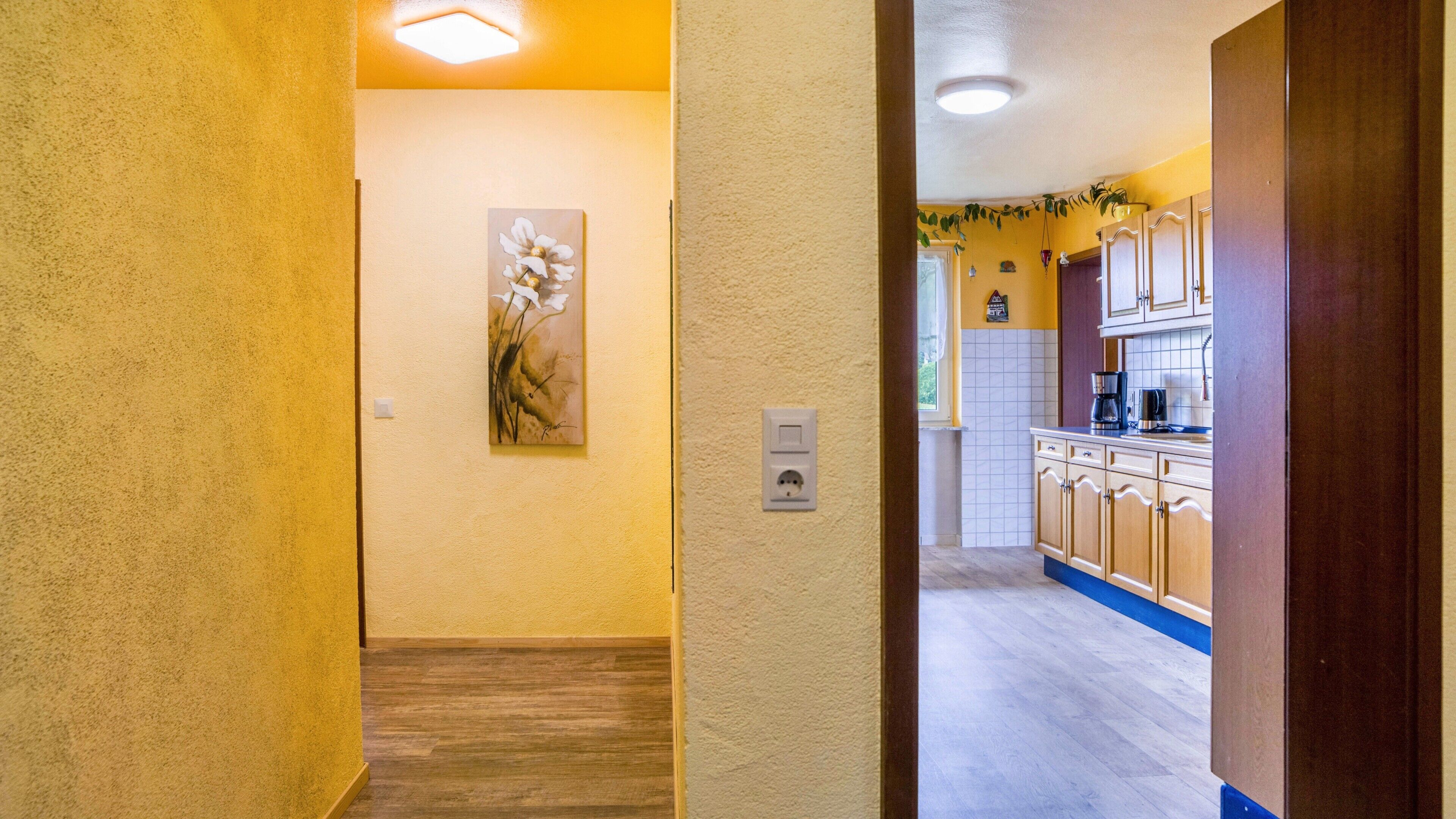 A hallway leading to a kitchen with wooden cabinets, a painting on the wall, and a light switch.