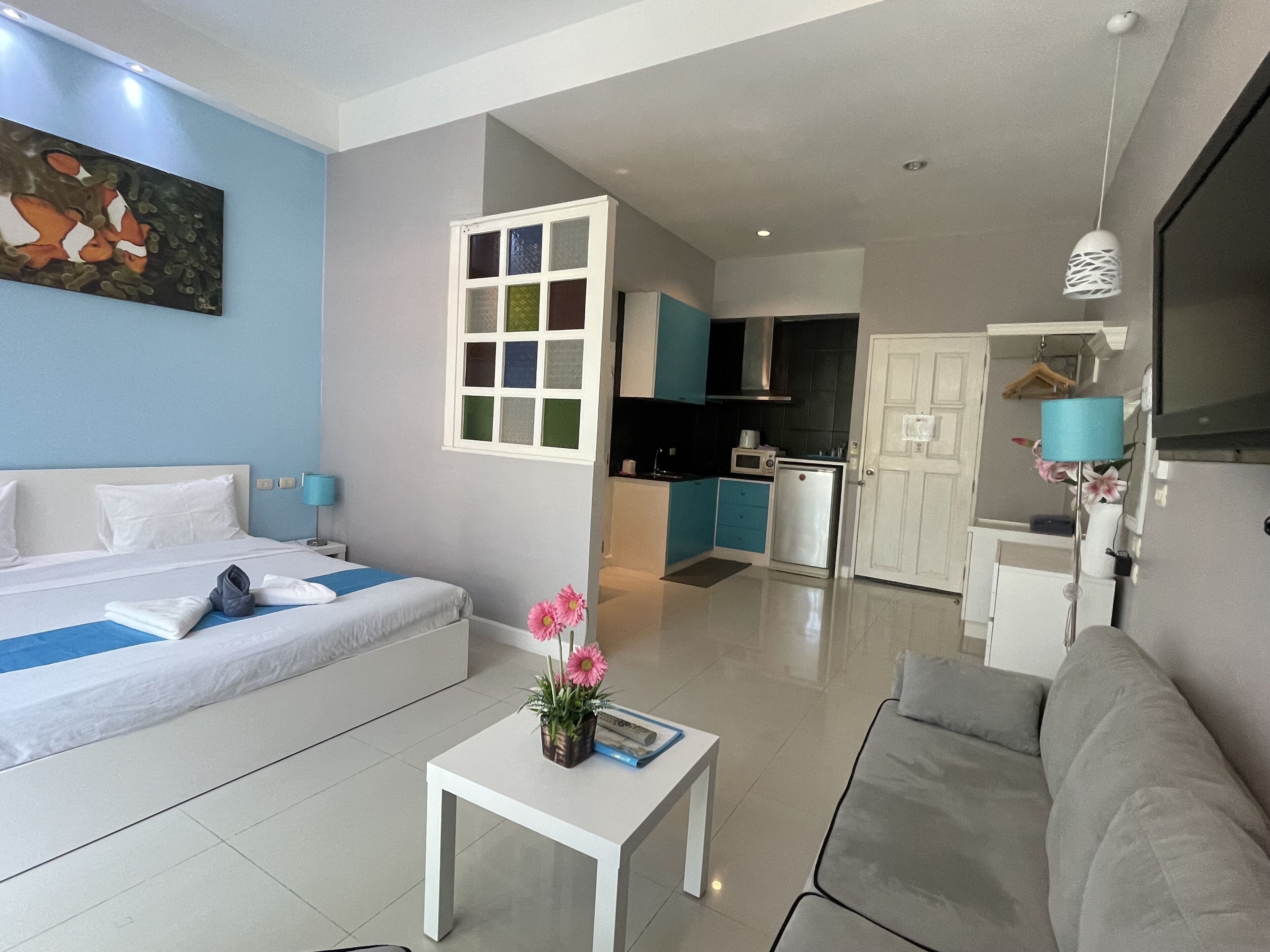 Une chambre d’hôtel moderne avec un lit, une petite table blanche ornée d’un vase de fleurs, un canapé gris et un coin cuisine visible à travers une porte ouverte.