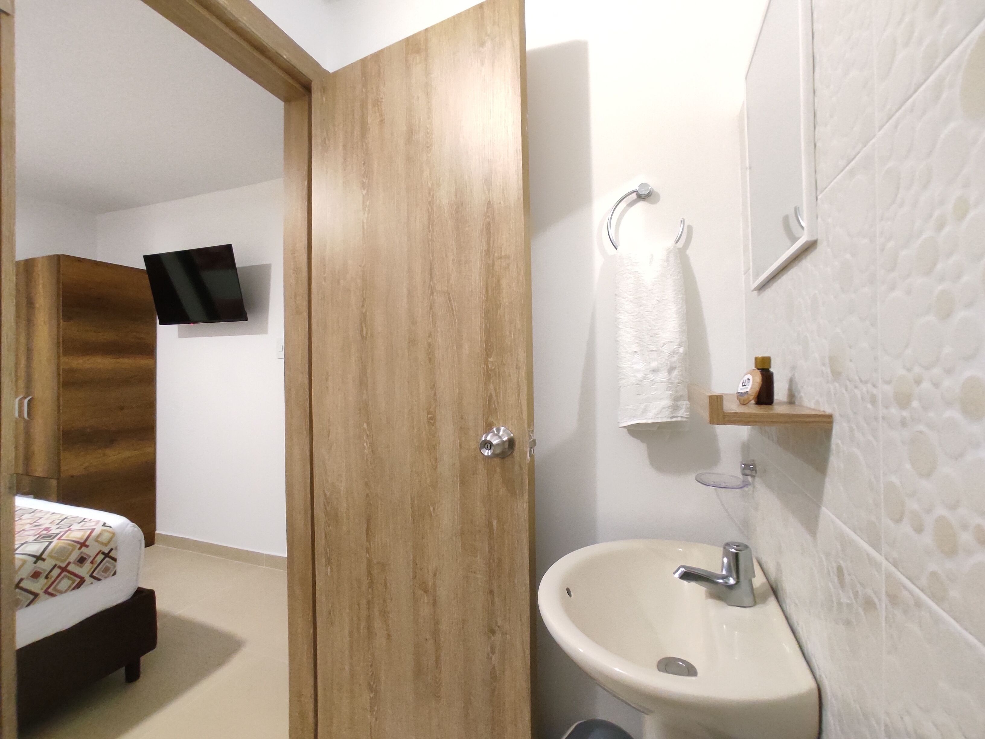 Habitación de hotel con armario de madera, cama con colcha estampada, televisor montado, baño con lavamanos, toalla y estante pequeño con artículos de aseo.
