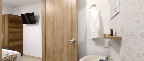 Habitación de hotel con armario de madera, cama con colcha estampada, televisor montado, baño con lavamanos, toalla y estante pequeño con artículos de aseo.
