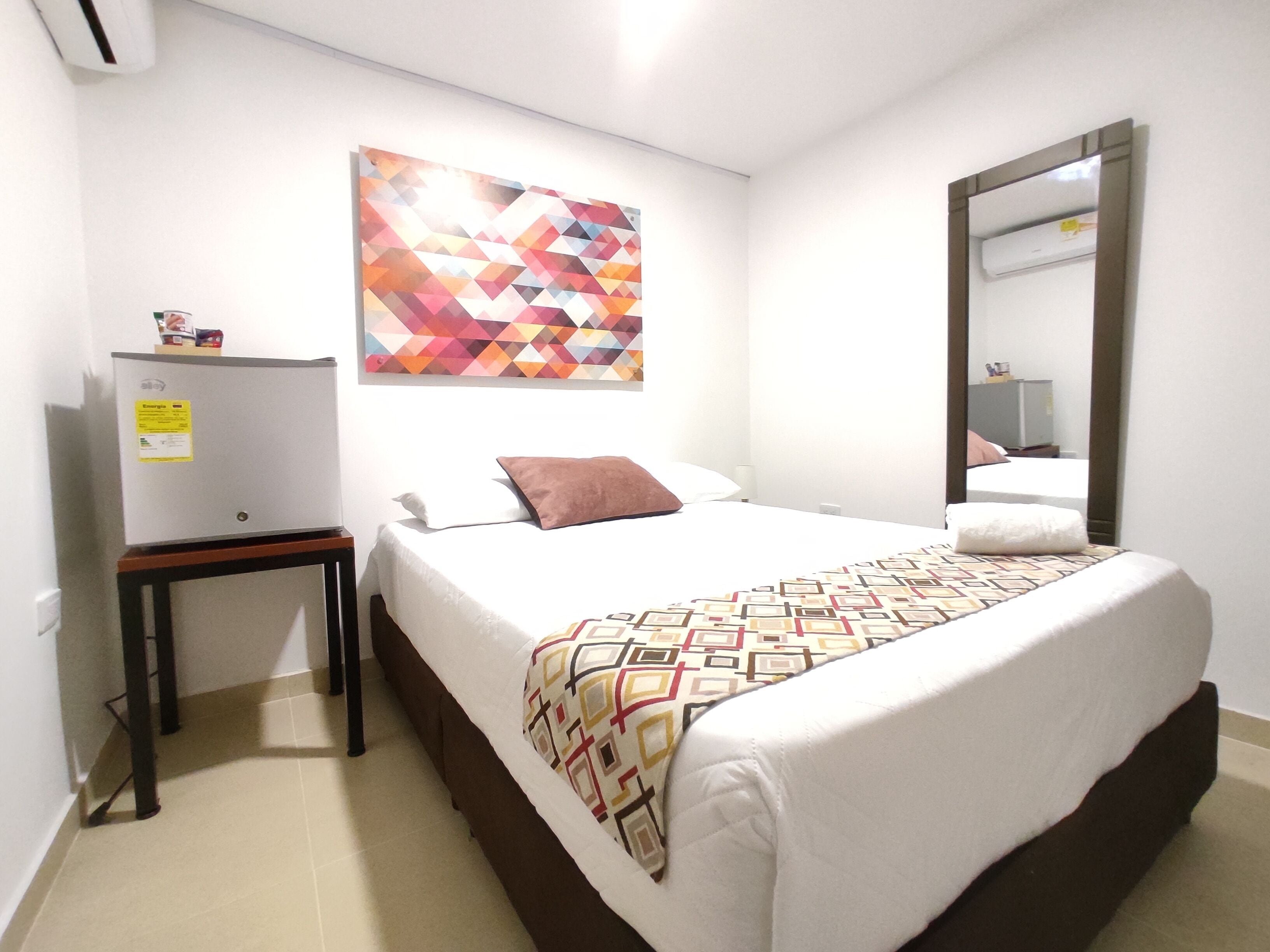 Habitación de hotel con cama, escritorio, espejo y un cuadro de arte geométrico y colorido.