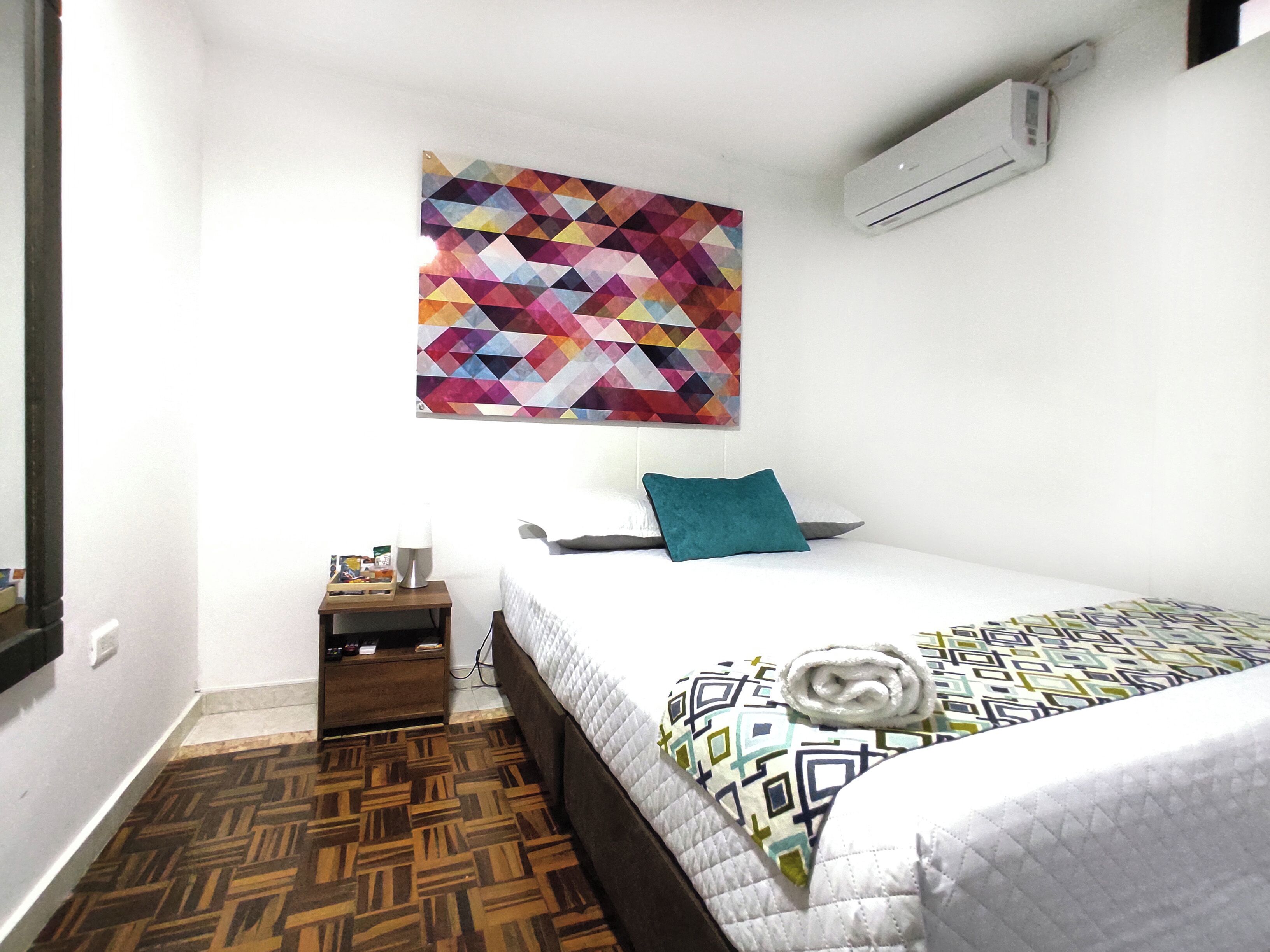 A bedroom with a bed, a nightstand, a wall-mounted air conditioner, and a geometric-patterned artwork.
