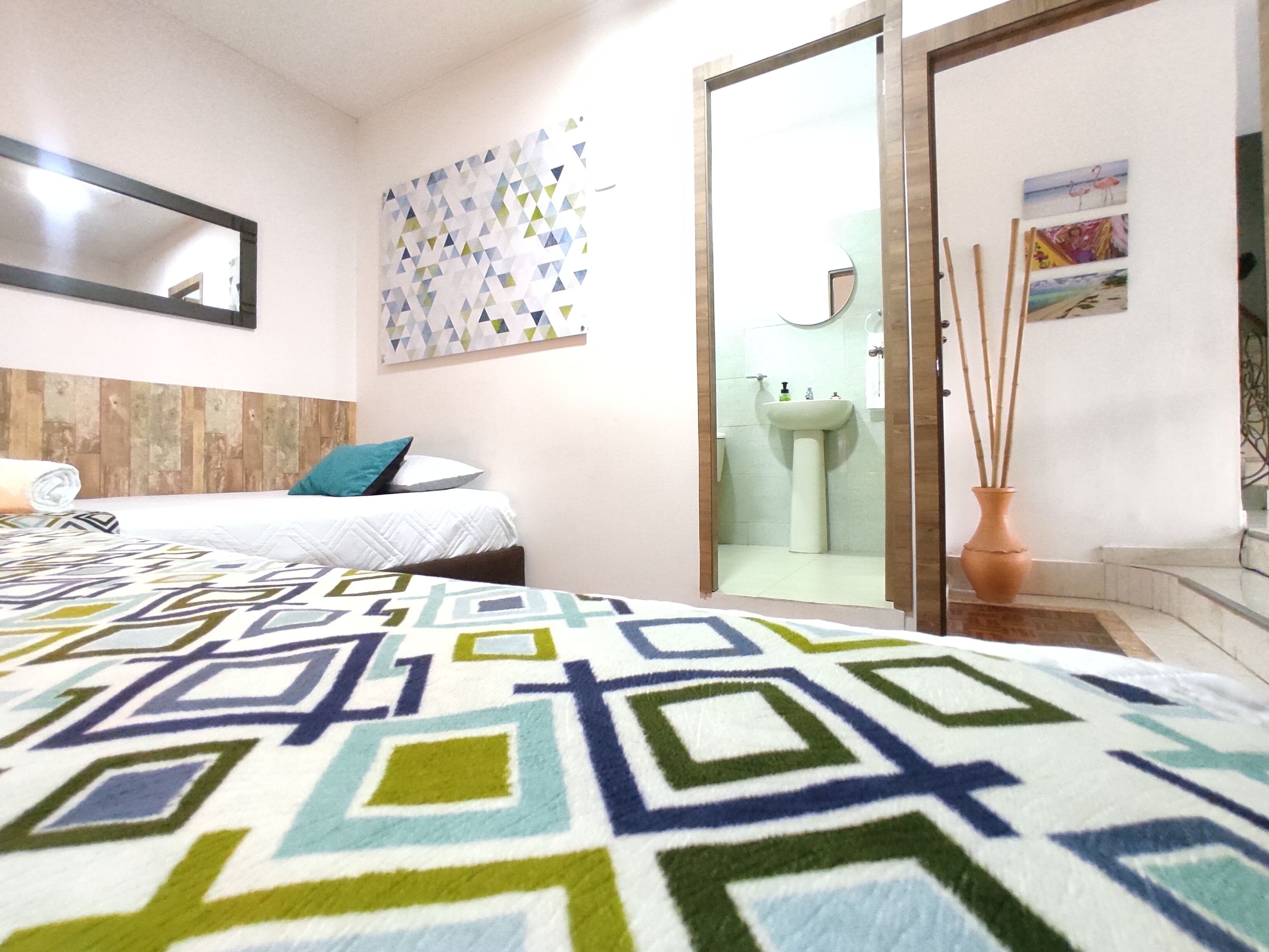 A hotel room with a bed, a bedside table, a mirror, a wall with a geometric-patterned artwork, a bathroom visible through an open door, and a potted plant.