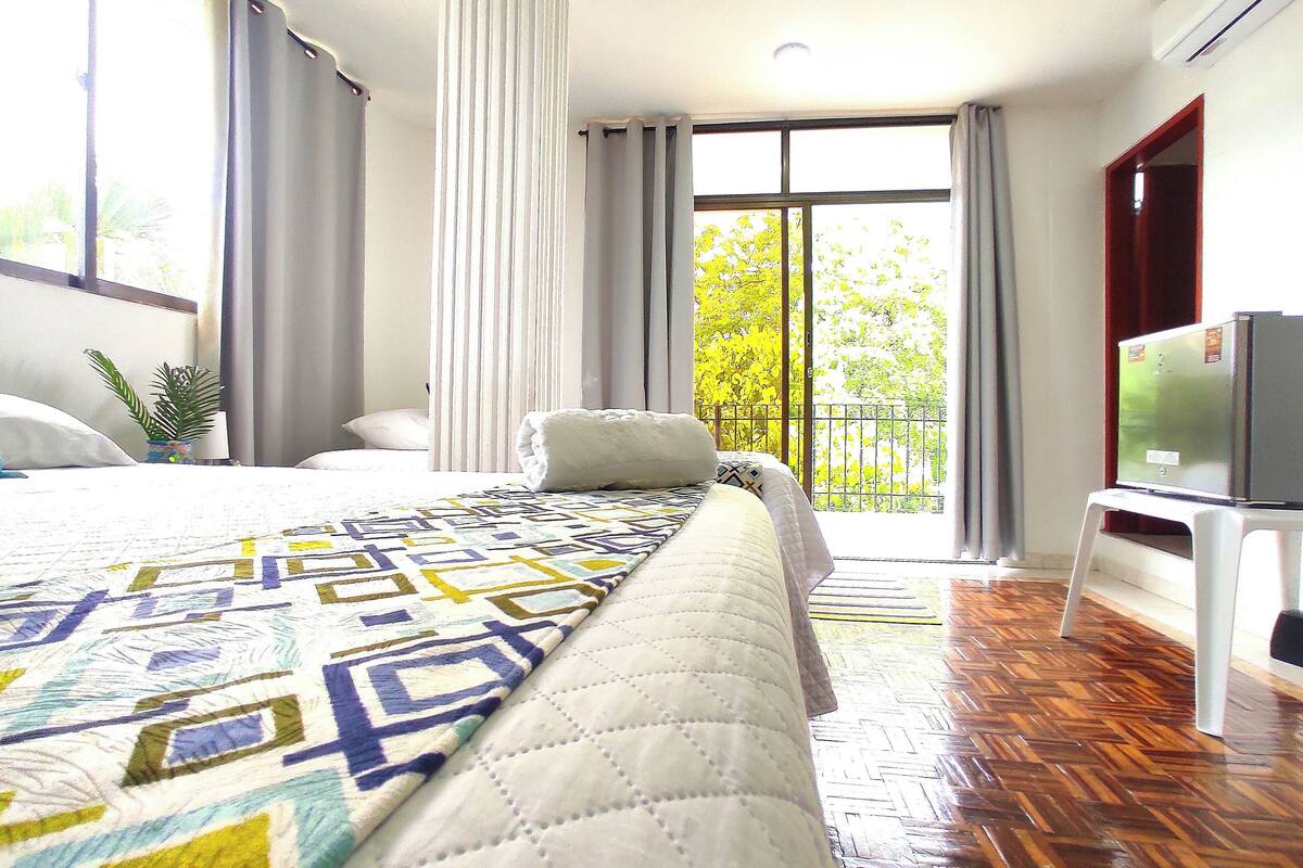 A hotel room with a large bed, a TV on a stand, a balcony with a view of trees, and a wooden floor.