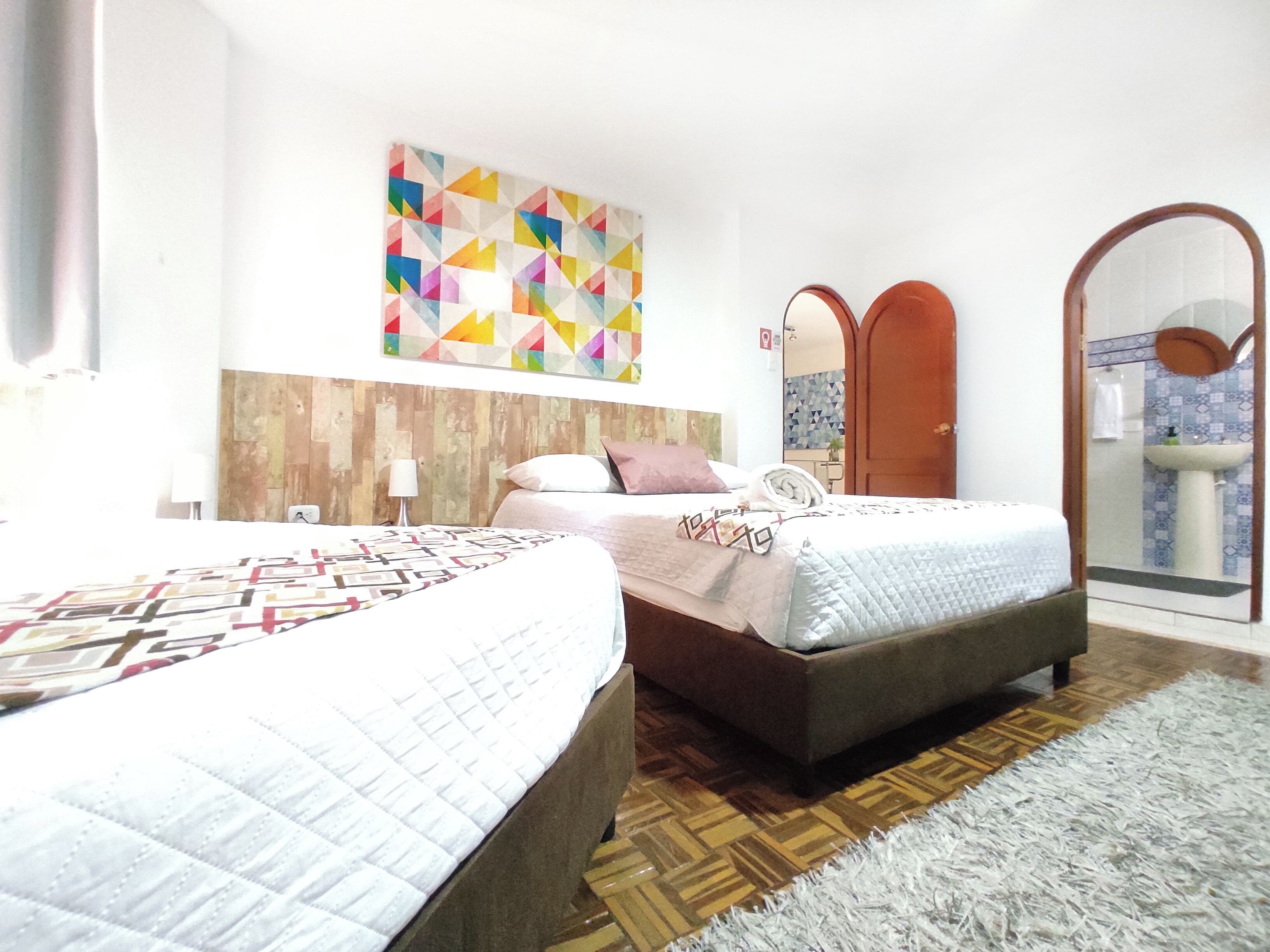 A hotel room with two beds, a wooden headboard, a colorful geometric wall art, a mirror, and a bathroom visible through an arched doorway.