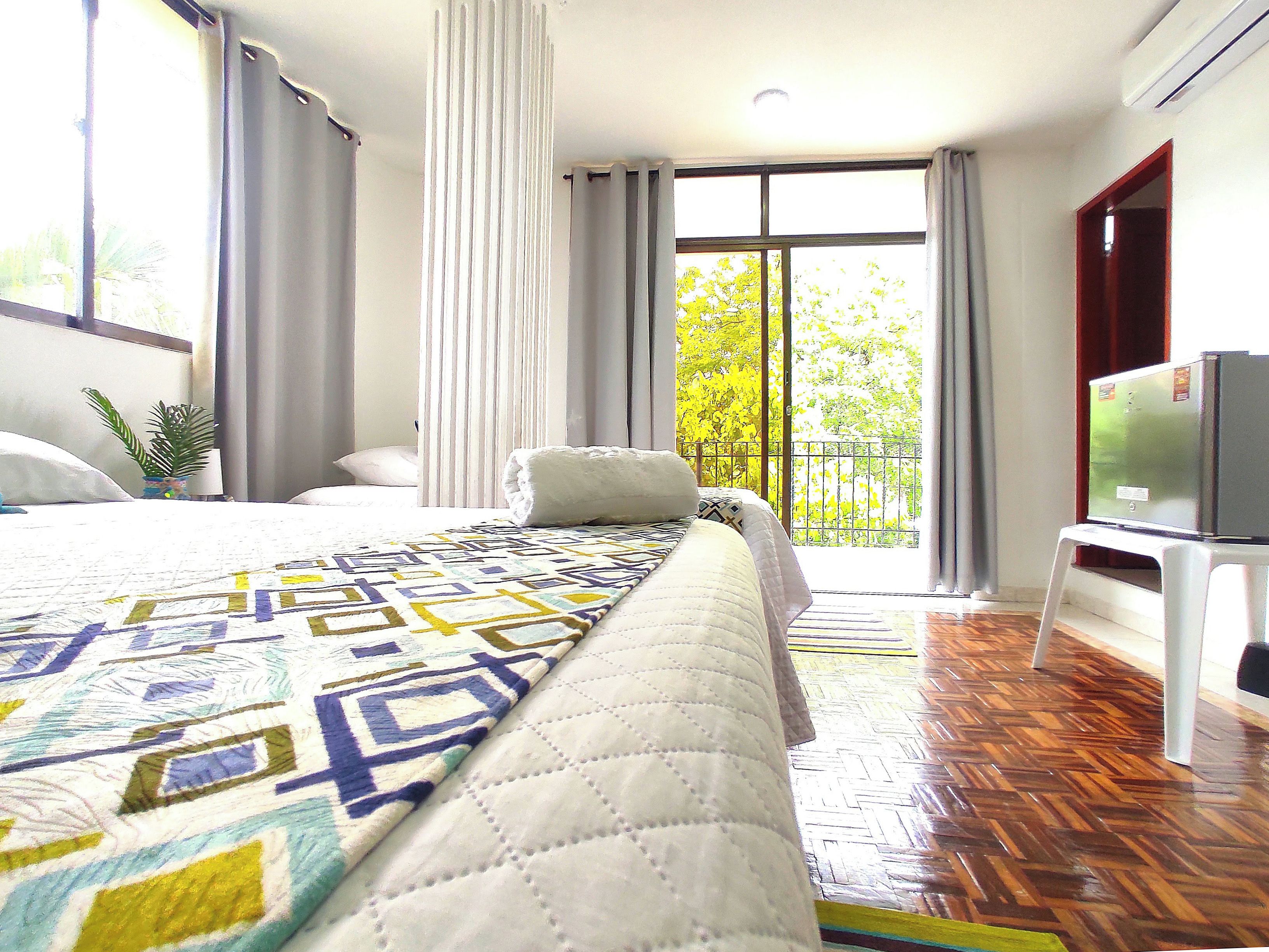 A hotel room with a large bed, a TV on a stand, a balcony with a view of trees, and a wooden floor.