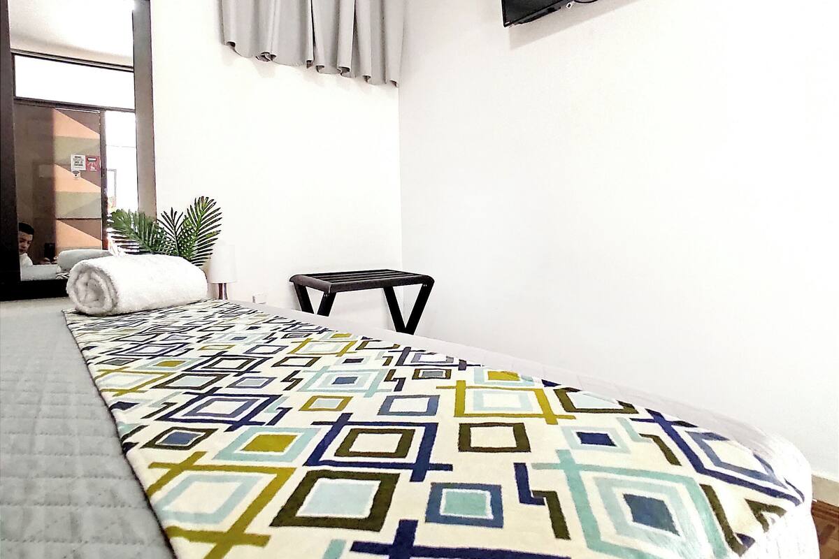A bedroom with a patterned bedspread, a flat-screen TV mounted on the wall, a window with curtains, and a small potted plant.