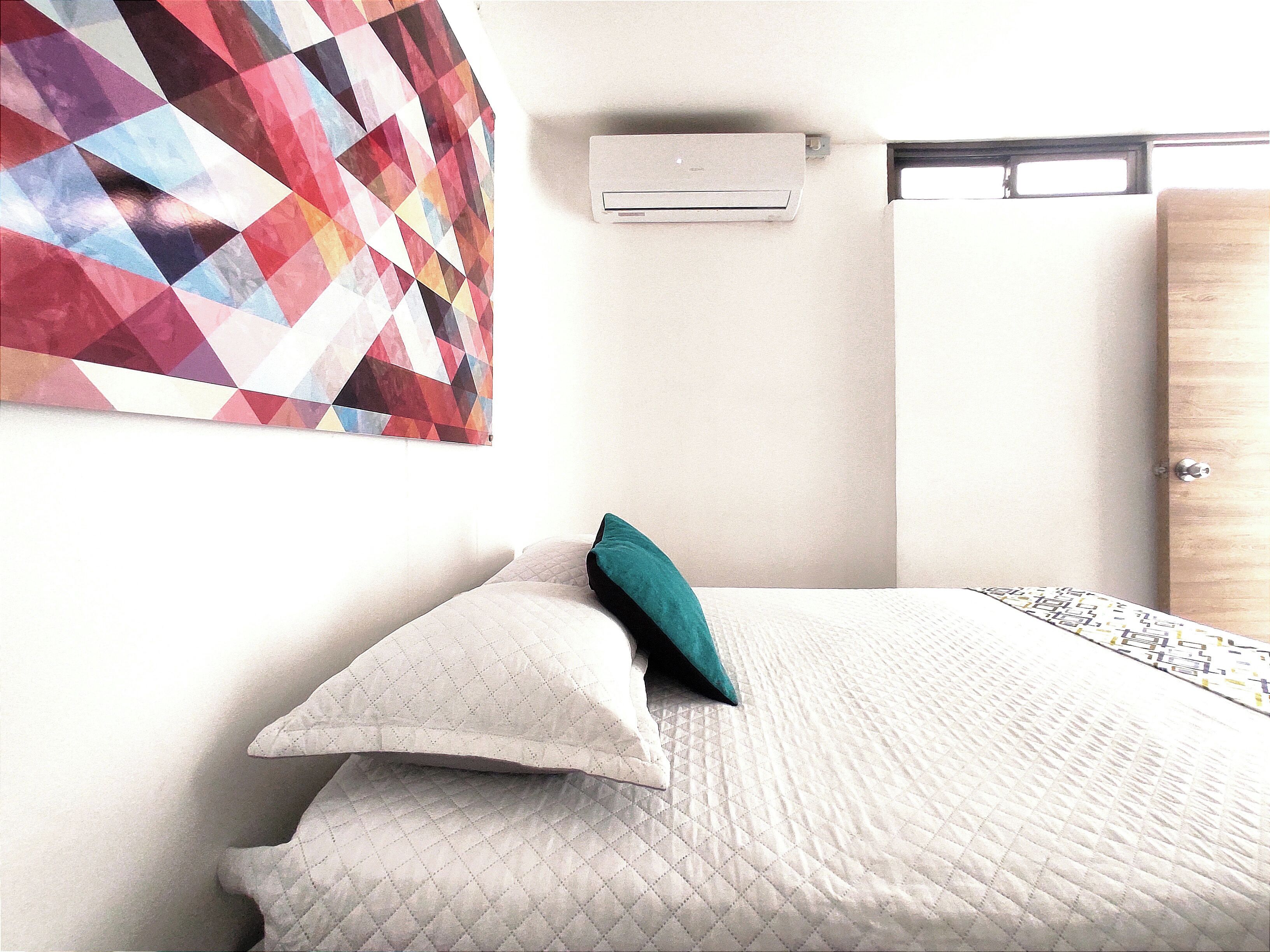 A bedroom with a bed, pillows, a quilted blanket, a wall-mounted air conditioner, and a geometric-patterned wall hanging.