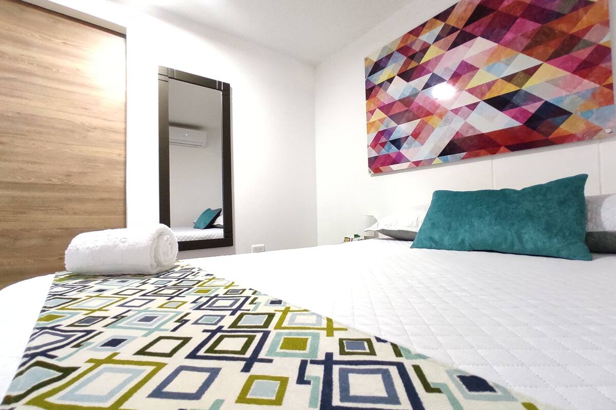 A hotel room with a bed, a colorful geometric wall art, a wooden door, and a white wall.