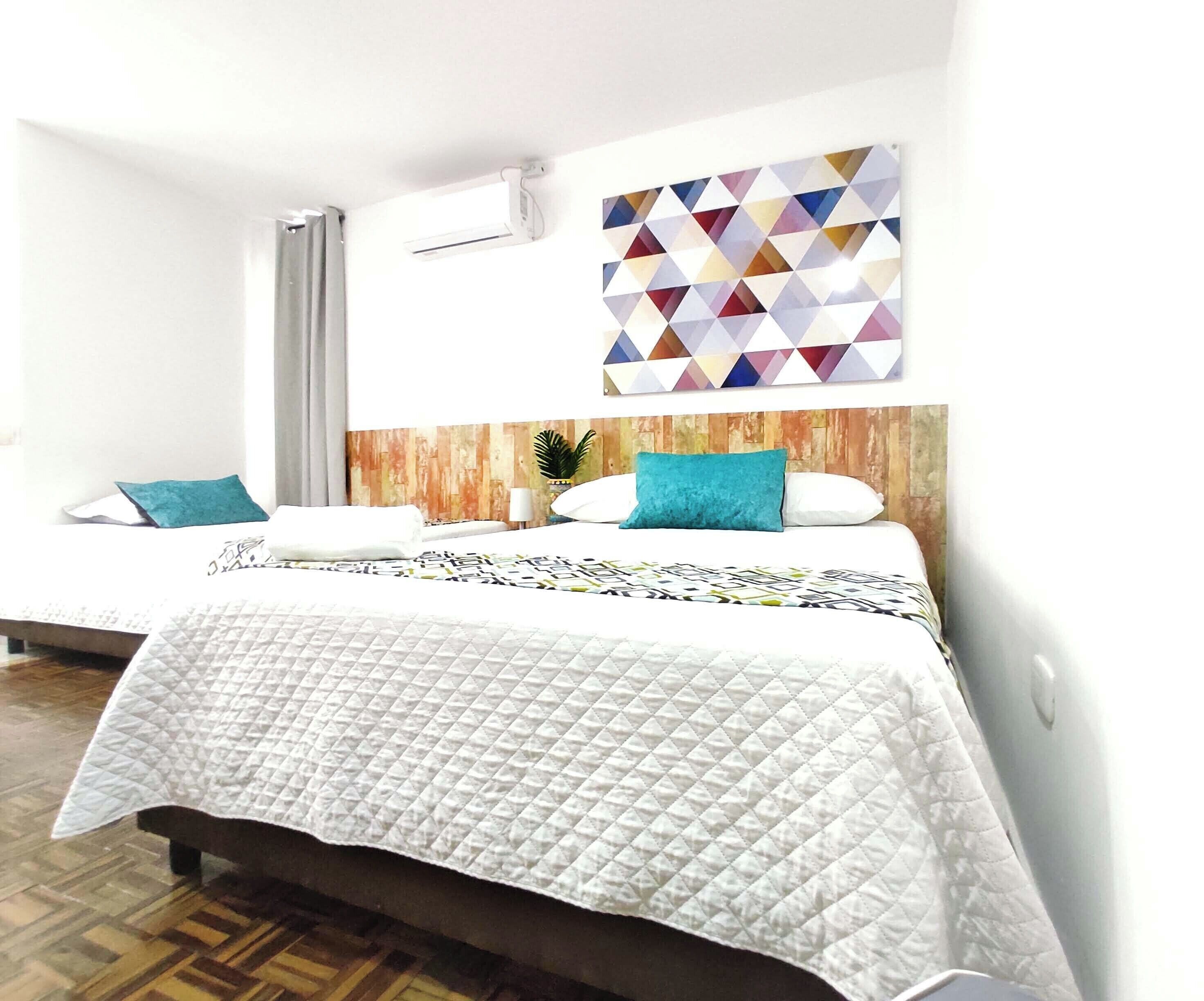 A hotel room with a bed, a wooden headboard, a geometric wall art piece, and a white air conditioning unit.