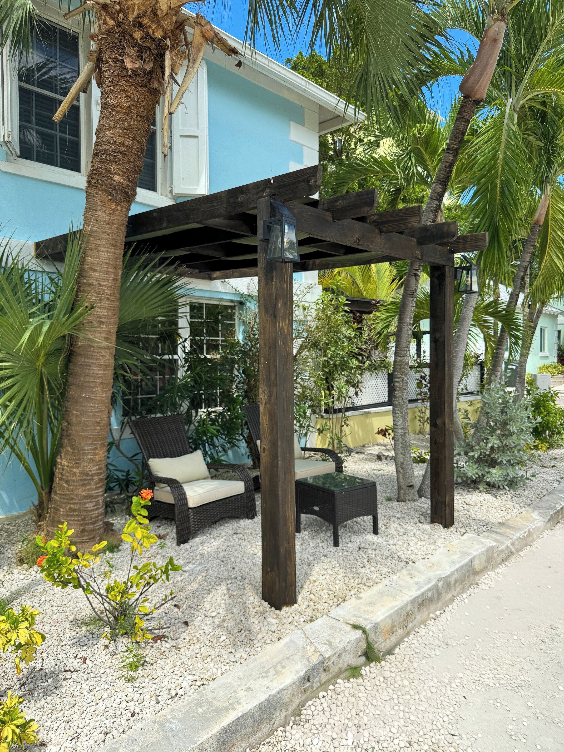 A covered outdoor seating area with wicker furniture, a palm tree, and a house in the background.