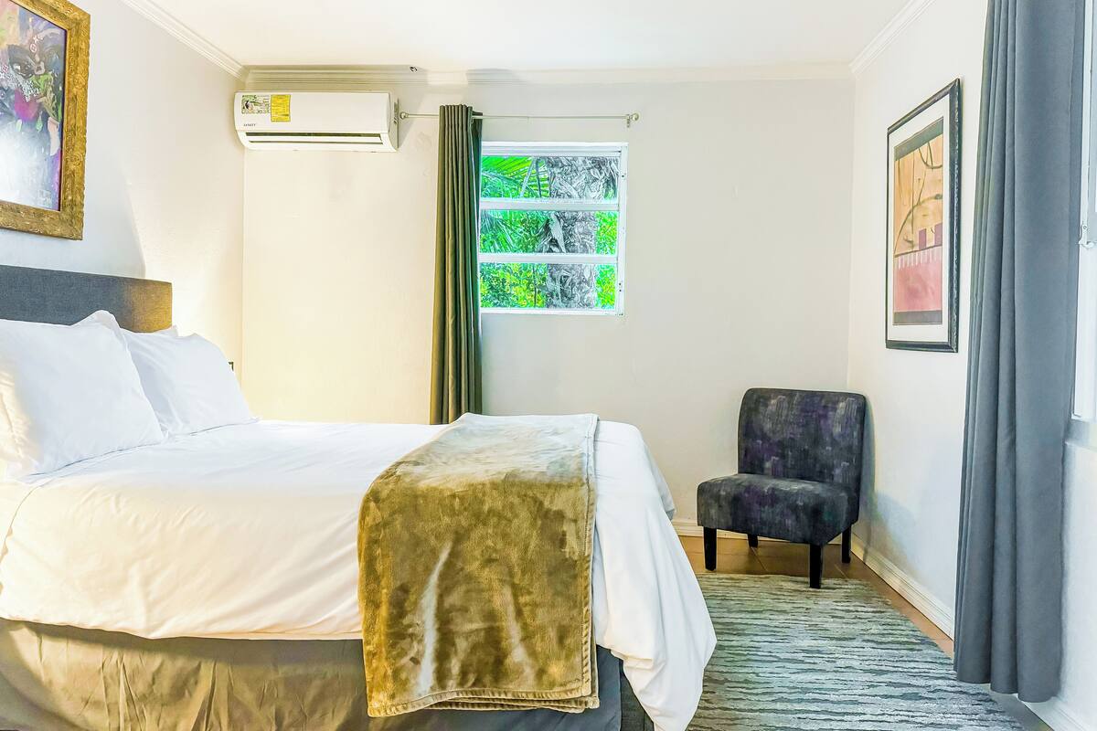 A hotel room with a bed, a chair, an air conditioning unit, a window with a view of greenery, and a ceiling fan.