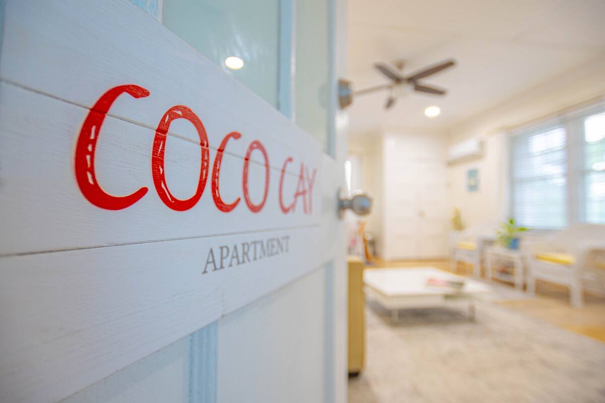 A sign for "Coco Cay Apartment" with a modern interior featuring a ceiling fan, a white sofa, and a coffee table.