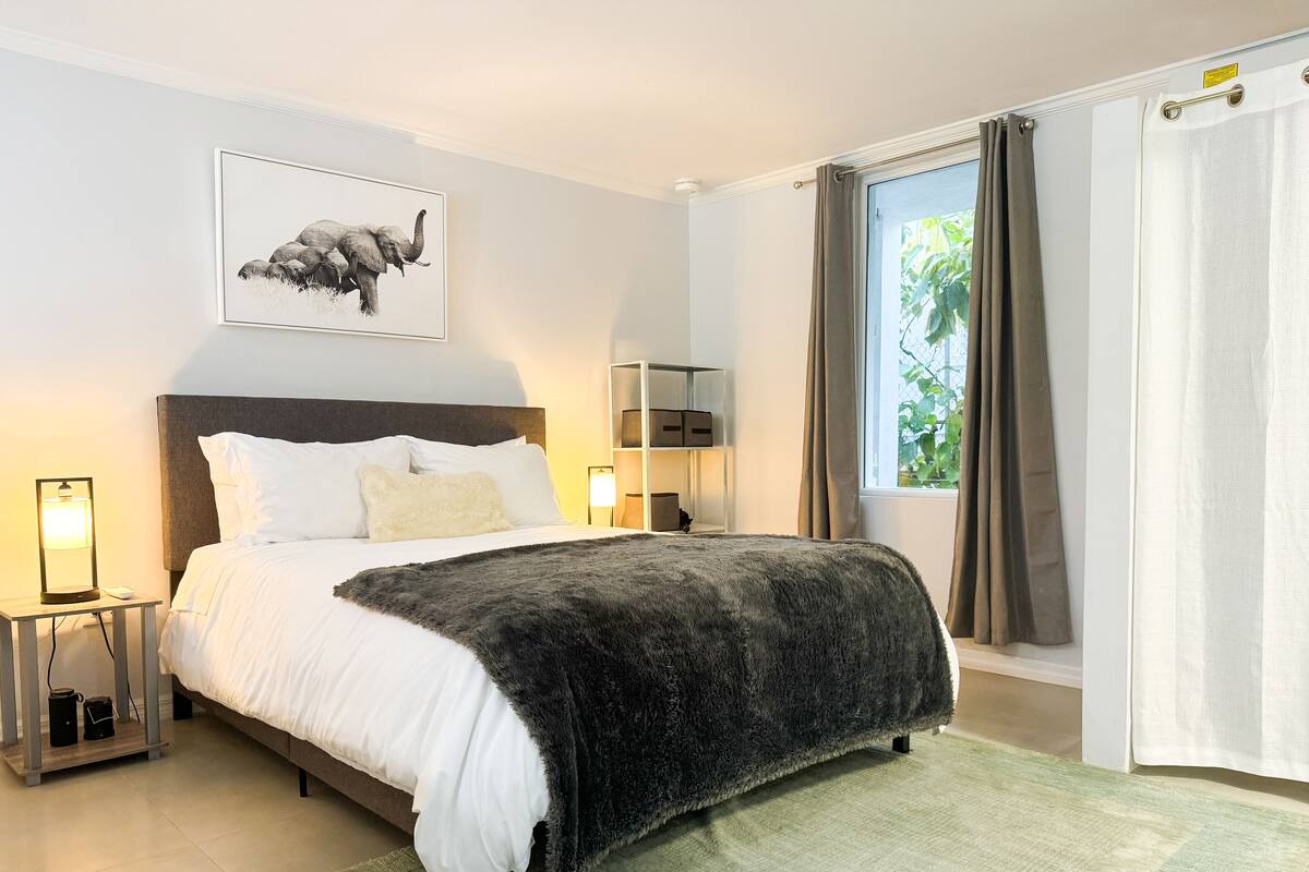 A modern bedroom with a large bed, a bedside table with a lamp, a framed picture on the wall, and a window with curtains.