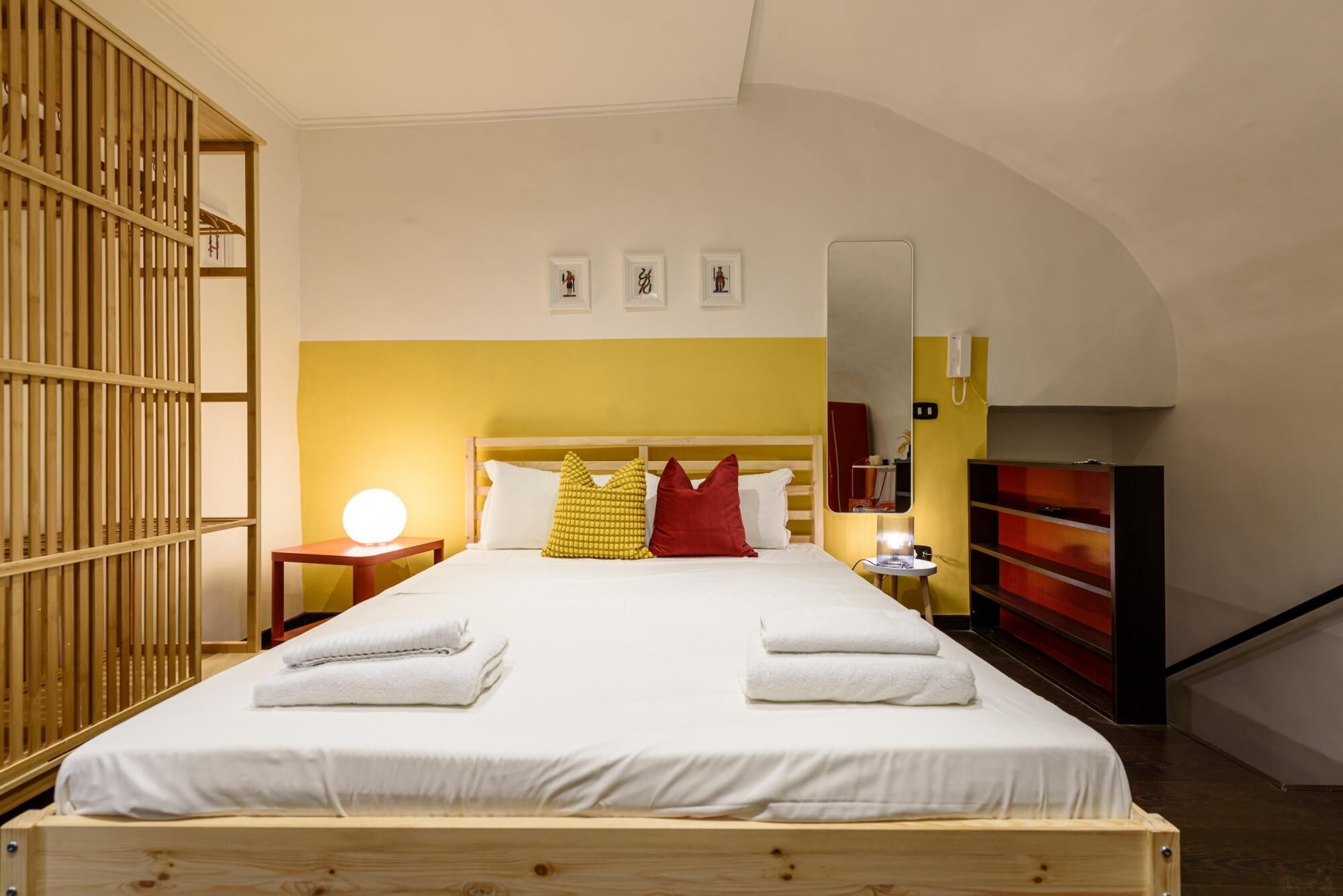 Ein Hotelzimmer mit einem hölzernen Bett, einem Nachttisch mit Lampe, einem Spiegel und einem roten Schrank.
