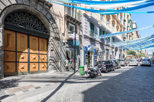 A cobblestone street with parked cars and a motorcycle, buildings with balconies, and a large wooden door. - Via Duomo 196 in Naples (Naples)