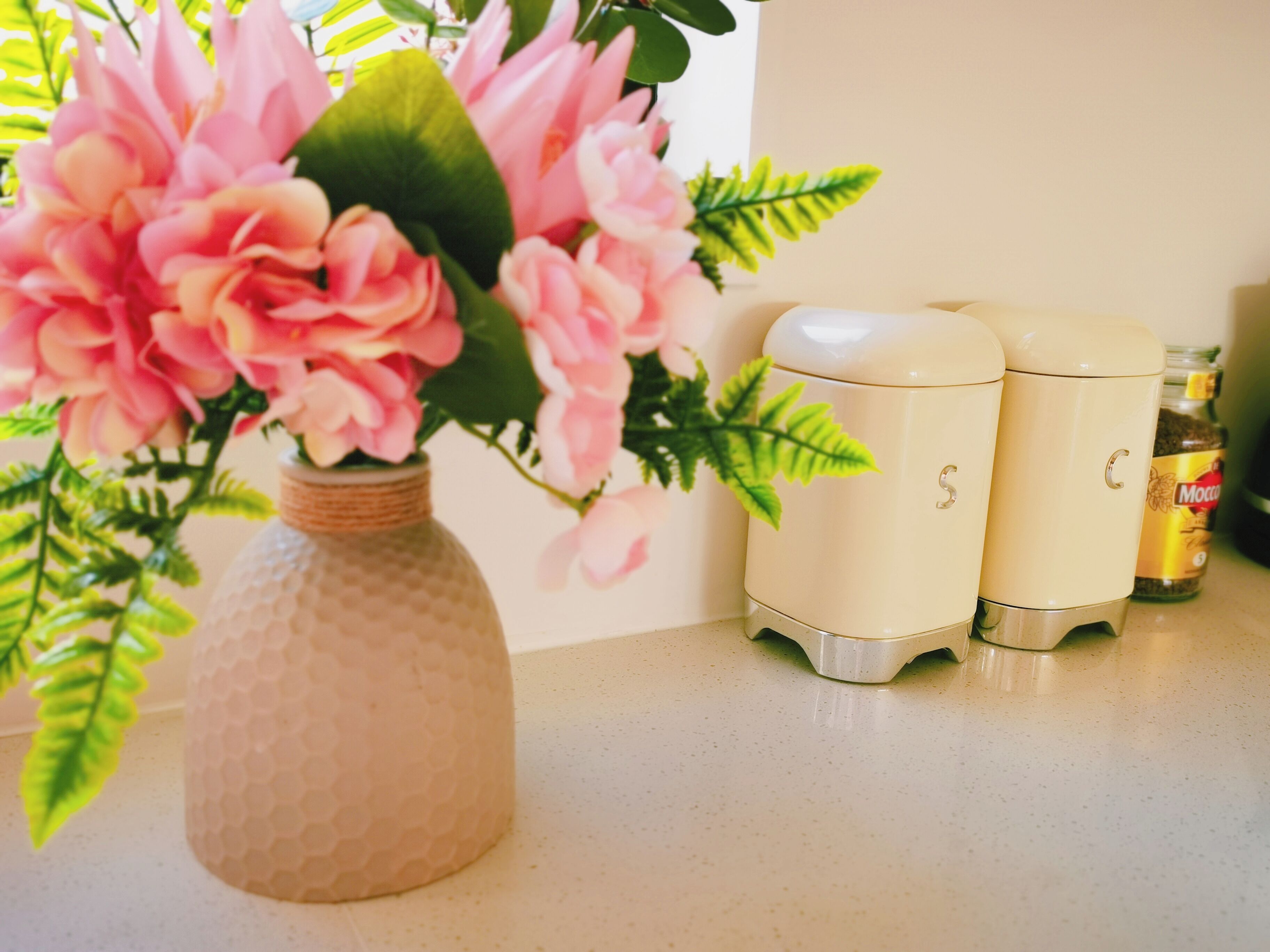 A kitchen counter with a decorative vase of pink flowers, a honeycomb-patterned ceramic container, and two cream-colored appliances.