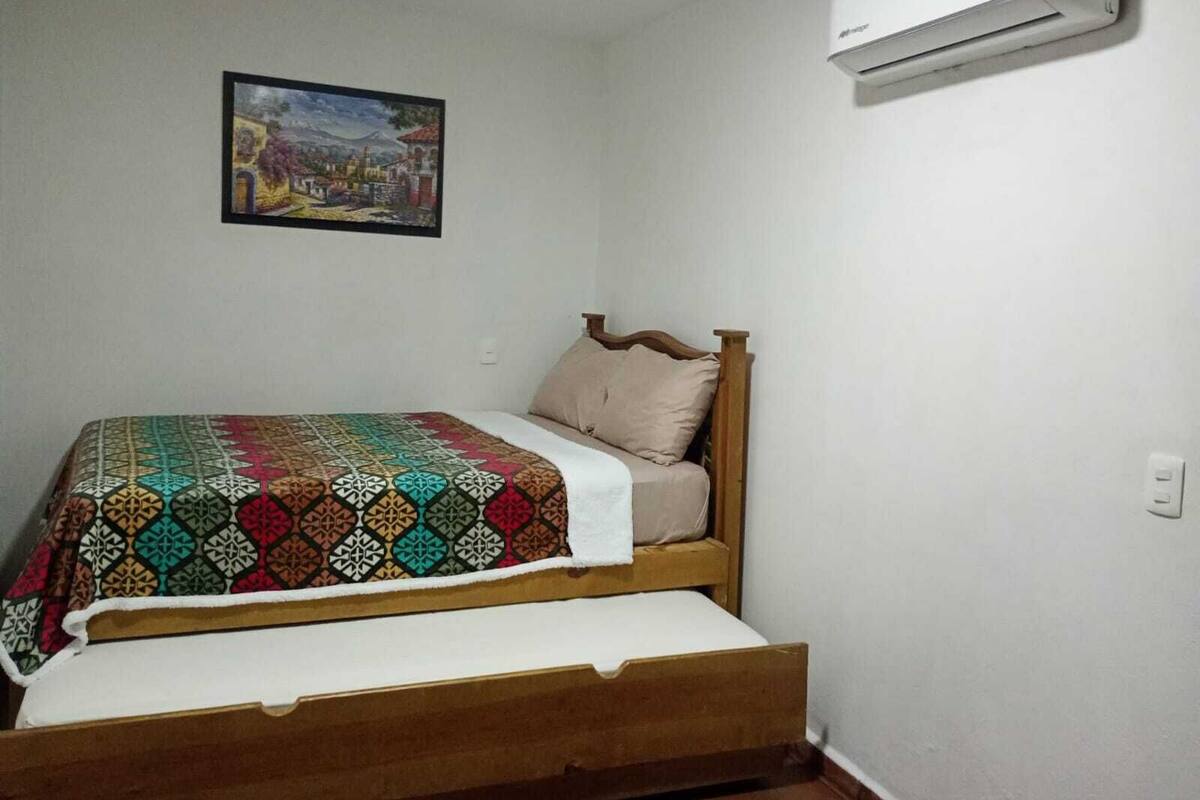 A single bed with a patterned blanket, a wooden headboard, and a wall-mounted air conditioner.
