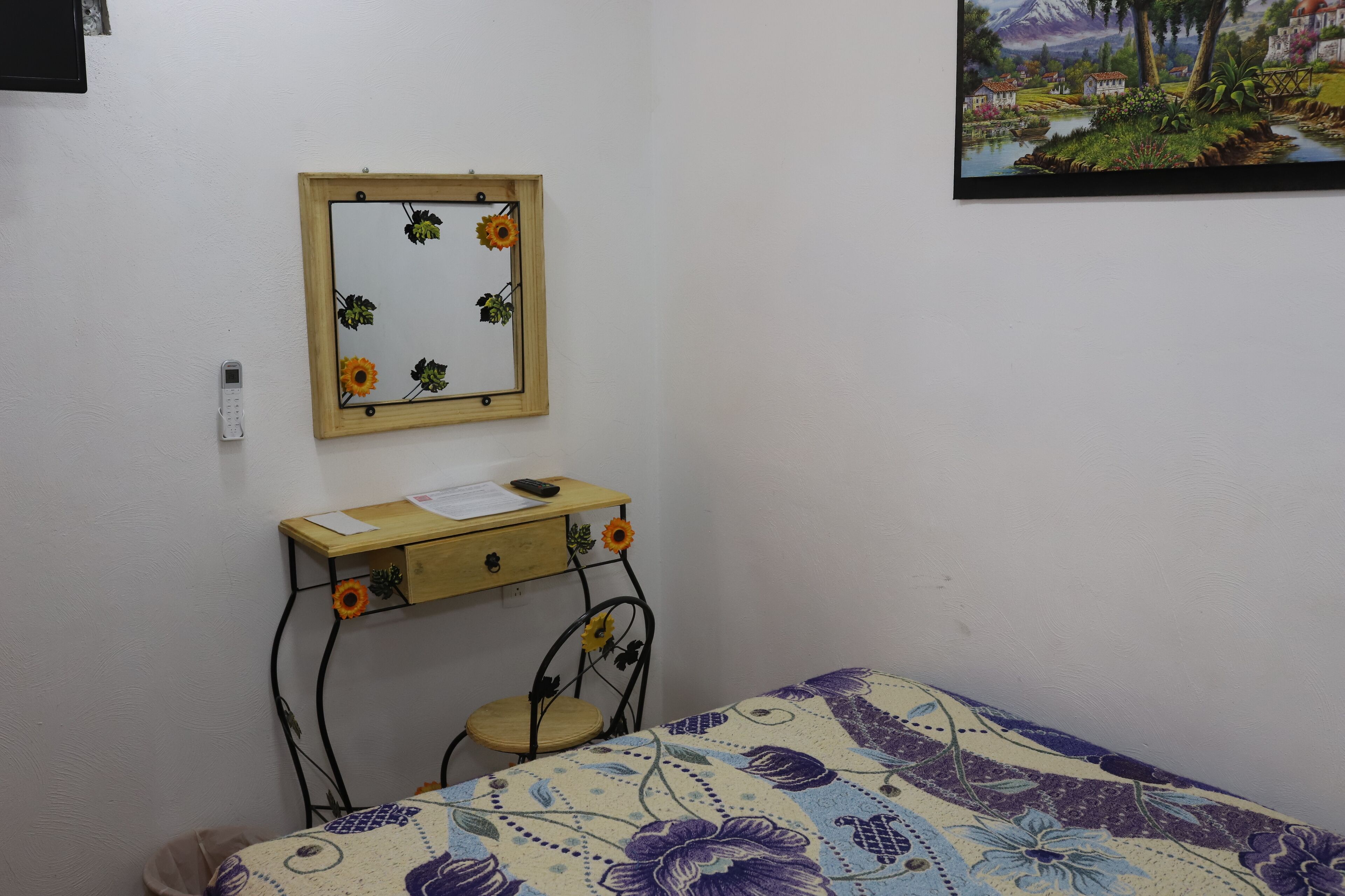 Un dormitorio con cama, escritorio con silla, espejo y un cuadro en la pared.