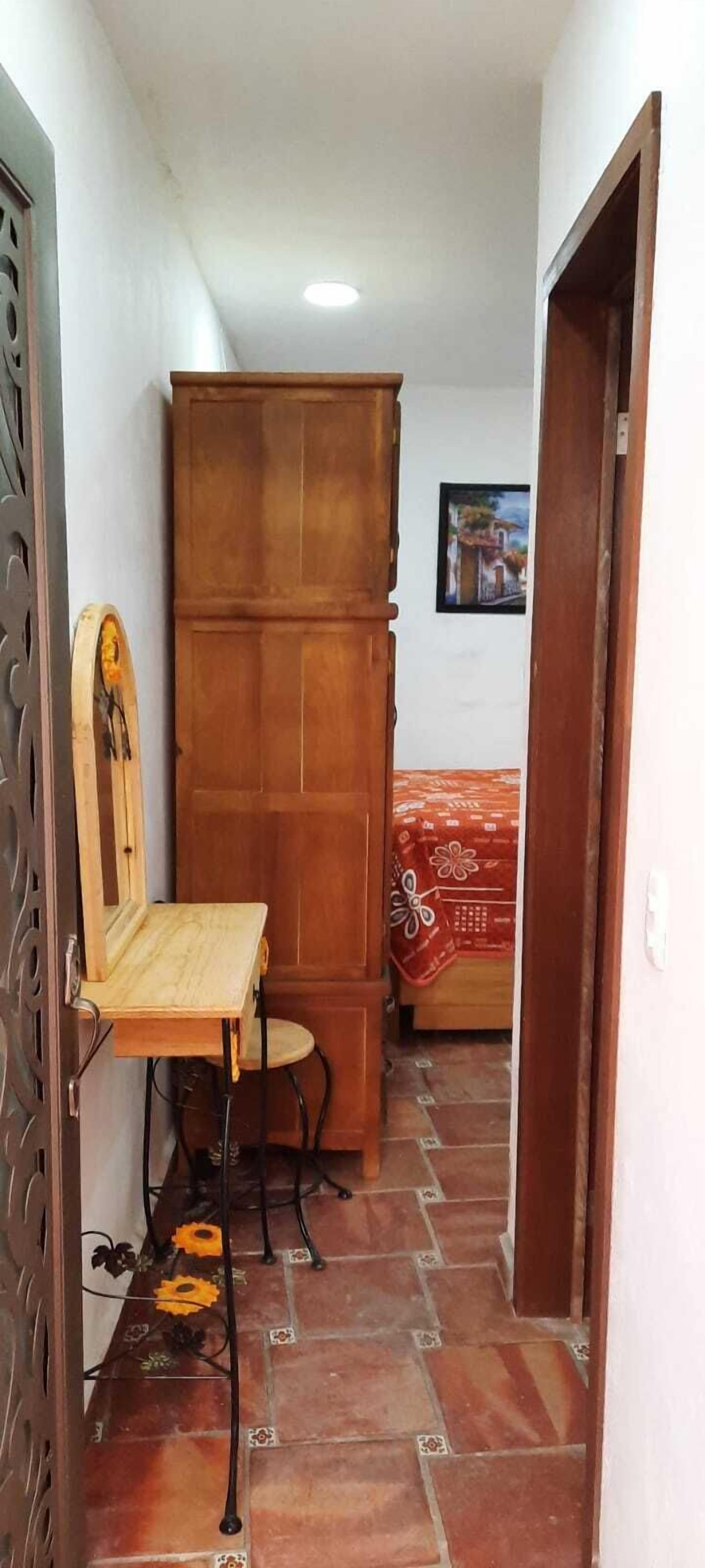 Ein schmaler Flur mit einem Holzschrank, einem Sessel und einem Bett mit gemustertem Überwurf.