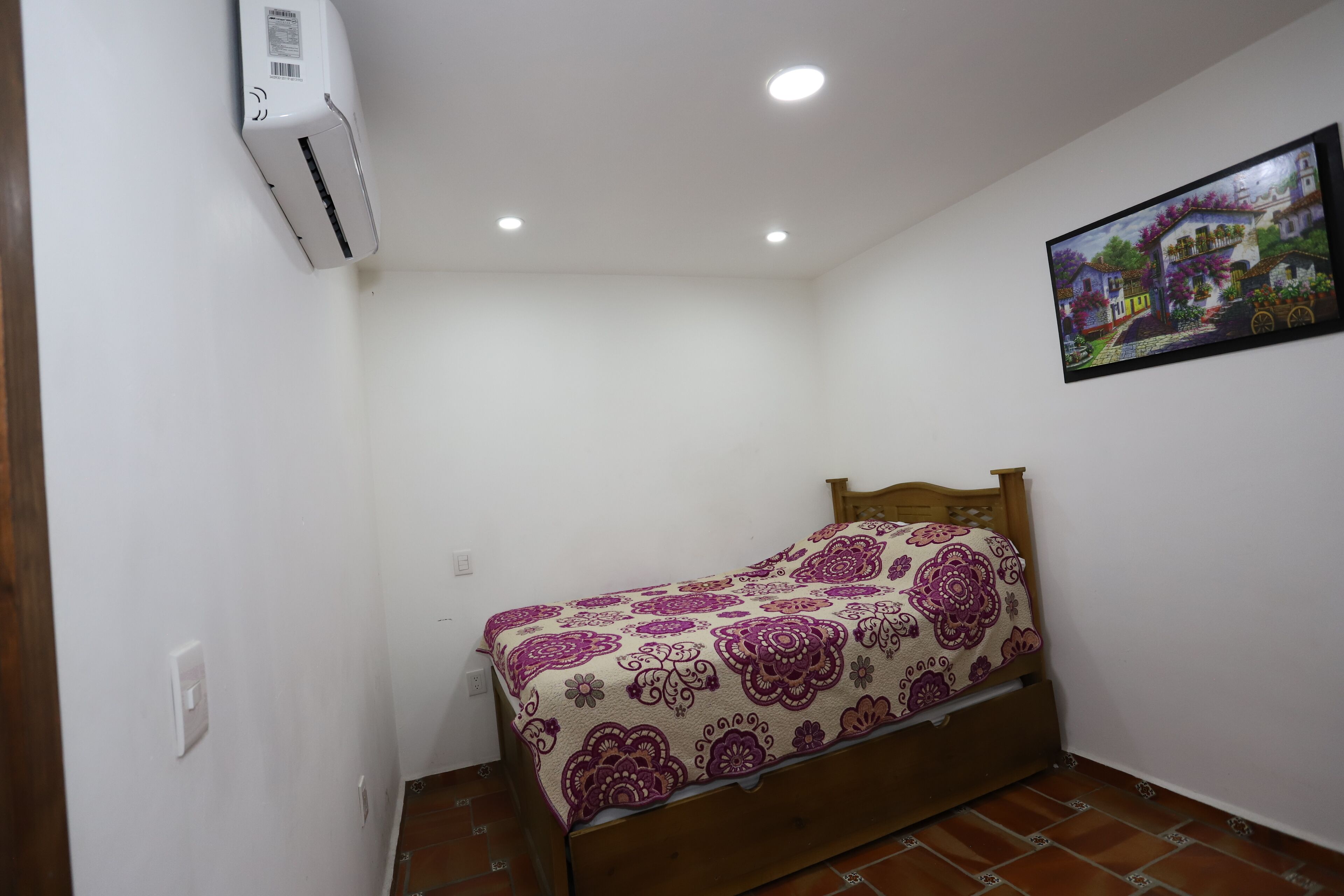 A small, single-bed room with a wooden headboard, a patterned bedspread, a wall-mounted air conditioner, and a framed picture on the wall.