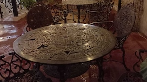Een terras buiten met een ronde metalen tafel en versierde stoelen, omgeven door bakstenen muren en hangende planten.