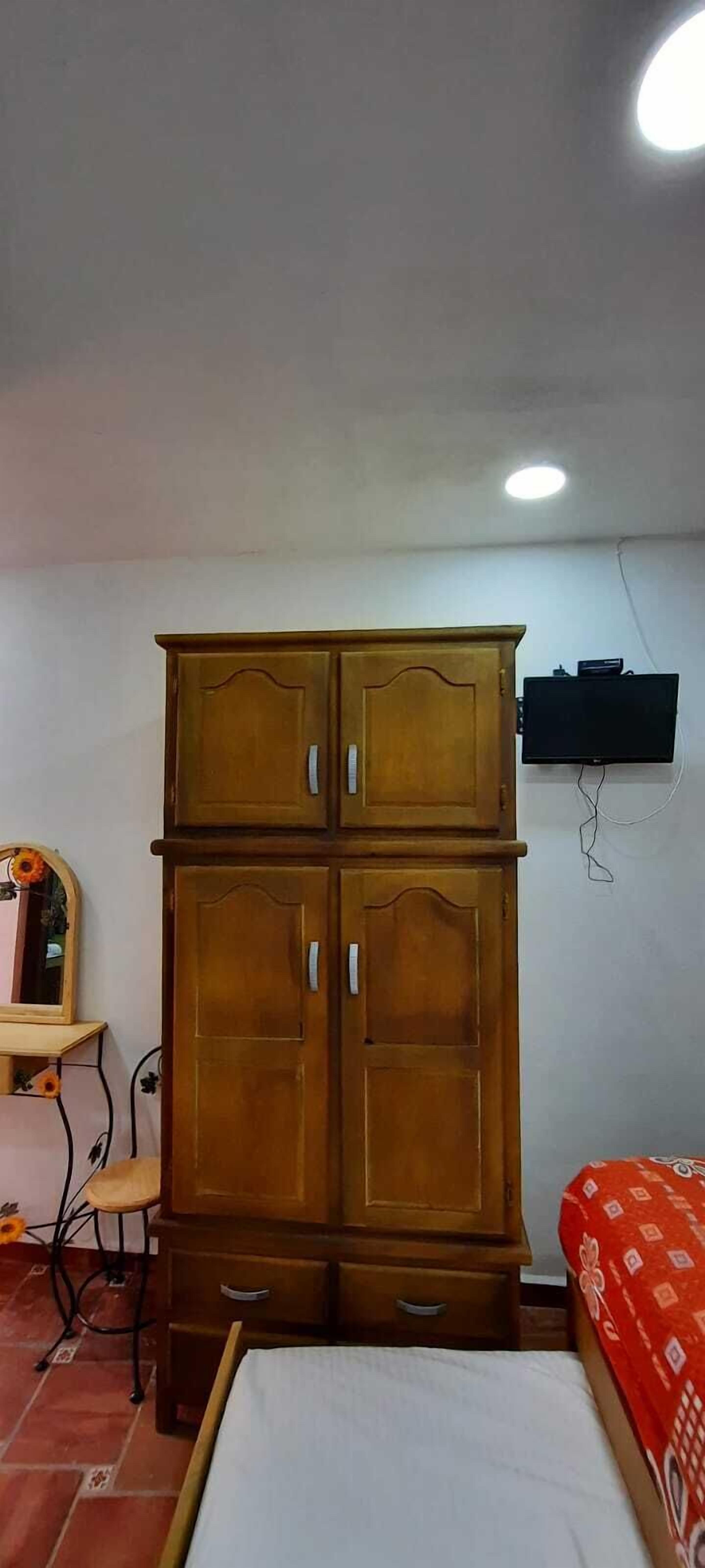 Una habitación con un armario de madera, una cama con colcha estampada, una silla, una mesita y un televisor montado en la pared.
