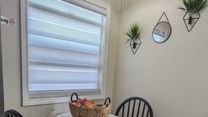 Un coin repas avec une table ronde, deux chaises noires, un panier tressé posé sur la table et une fenêtre avec des stores.