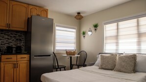 Une chambre à coucher de taille réduite, comprenant un lit, des tables de chevet, une petite kitchenette et une fenêtre avec des stores.
