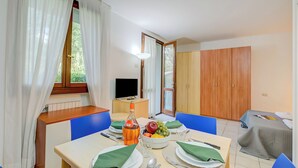 Un coin repas avec une table dressée pour deux personnes, un panier de fruits, une bouteille et un téléviseur en arrière-plan.