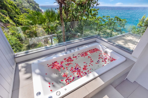 A modern bathroom with a bathtub filled with rose petals, overlooking a coastal view.