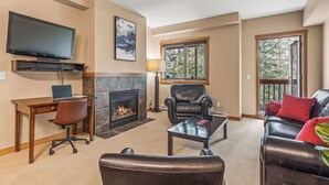 A living room with a fireplace, a television mounted above it, a leather sofa, a desk with a laptop, and a window with a view of trees. - Spacious King Bed with Pool & Hot Tubs (Canmore)