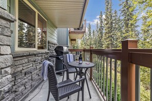 A balcony with wicker furniture, a grill, and a bottle of wine. - Spacious King Bed with Pool & Hot Tubs (Canmore)