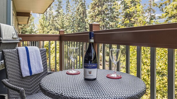 A wicker chair with a towel, a bottle of Ruffino Chianti wine, and two wine glasses on a round table on a balcony with a railing and trees in the background. - Spacious King Bed with Pool & Hot Tubs (Canmore)