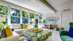 A spacious living area with a sofa, a coffee table with a tropical leaf pattern, a television, and a view of palm trees through large windows. - Vahevala: Your Tranquil Mid-Town Key West Oasis, 3BR Home, Pool & Close to Beaches & Duval Street! (Key West)