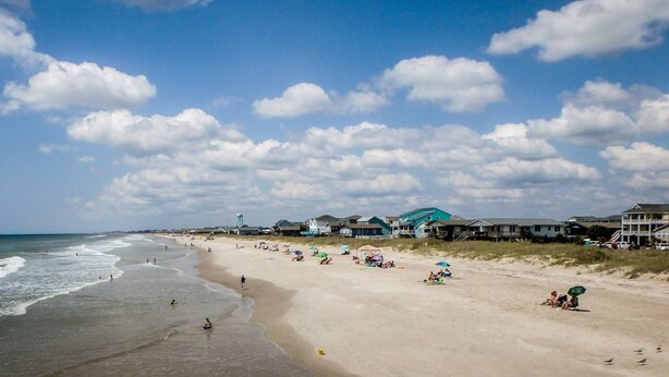 A beach with houses in the background, a clear sky, and people on the sand.
